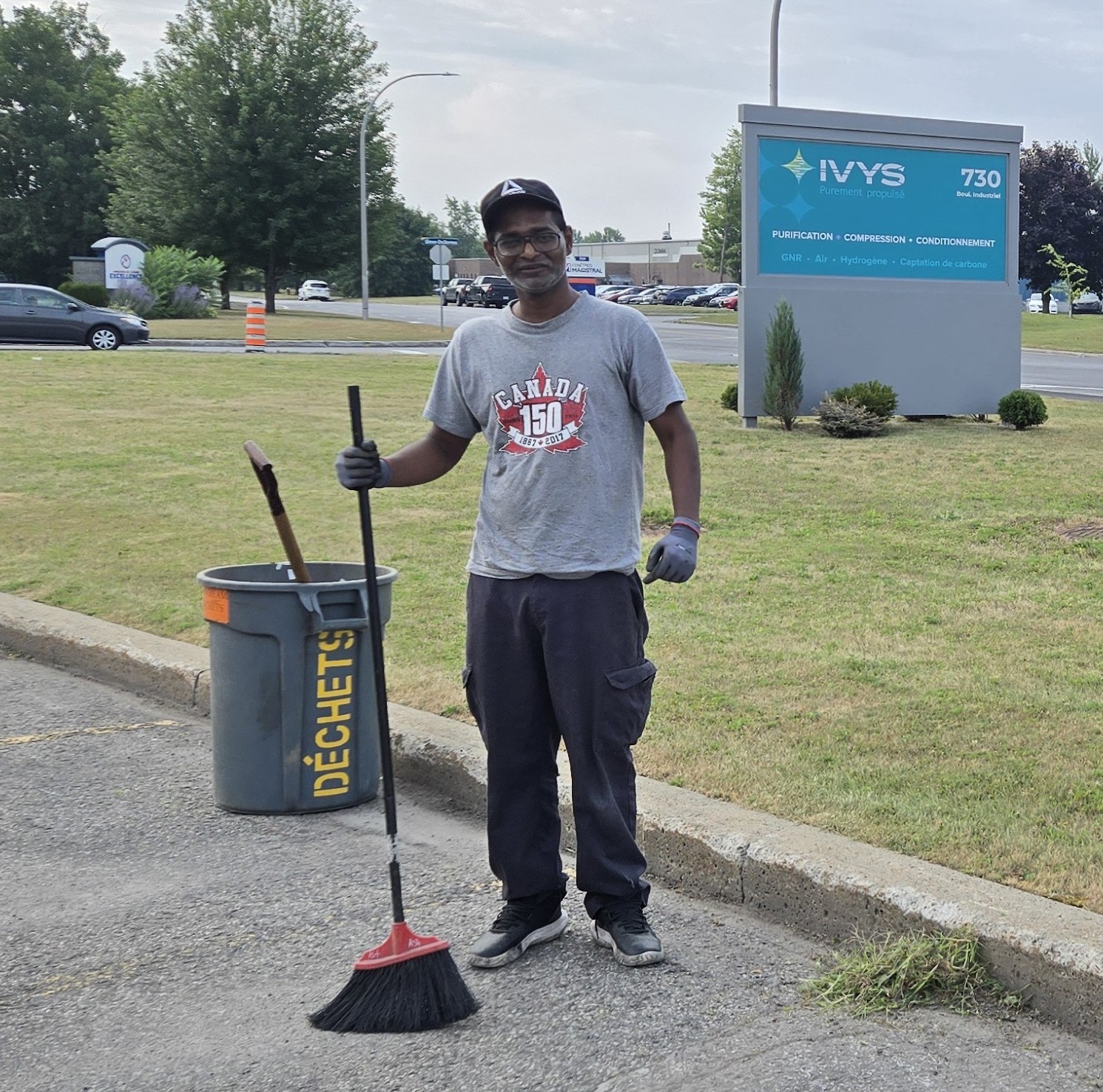 Thanushan, assembleur électrique chez Biostream, debout devant l’enseigne extérieure d’Ivys, tenant un balai à la main. Il s’est porté volontaire pour nettoyer le stationnement. Il a une expression de fierté.
