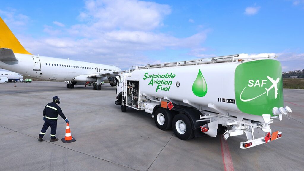 SAF-feuling-cut une scène sur le tarmac d’un aéroport, où un camion-citerne de carburant est stationné à proximité d’un avion. Le camion est clairement identifié comme transportant du carburant d’aviation durable, avec l’inscription « Sustainable Aviation Fuel » (SAF) bien visible sur le flanc, accompagnée d’un symbole de goutte verte, représentant l’aspect écologique du carburant. Un travailleur en tenue de sécurité — gilet haute visibilité et casque — est en train de placer ou retirer un cône de signalisation orange près du camion, ce qui indique une opération en cours ou une mesure de sécurité. À l’arrière-plan, on aperçoit un avion stationné.