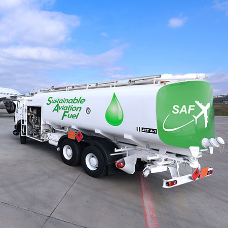 A fuel tanker truck parked on an airport tarmac. This vehicle is clearly marked as carrying sustainable aviation fuel, with the words “Sustainable Aviation Fuel” clearly visible on the side. A green drop symbol accompanies the text, highlighting the fuel’s eco-friendliness. On the rear of the truck, the acronym “SAF” (for Sustainable Aviation Fuel) is displayed alongside a graphic of an airplane. The truck is positioned in a typical airport environment, with an asphalt surface and infrastructure elements visible in the background.